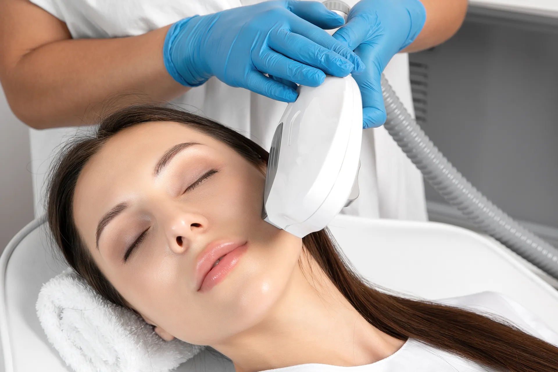Young woman receiving a facial treatment with a laser device by a cosmetologist wearing blue gloves.