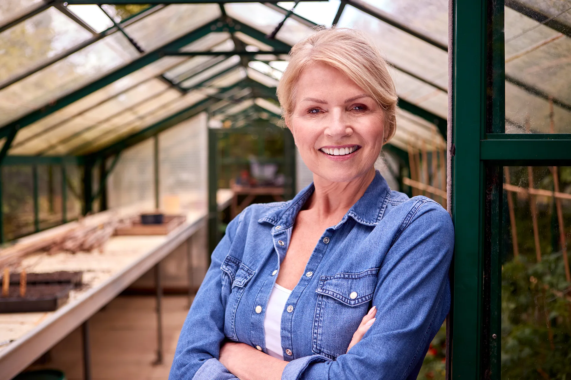 Smiling woman in a denim shirt standing in a greenhouse.