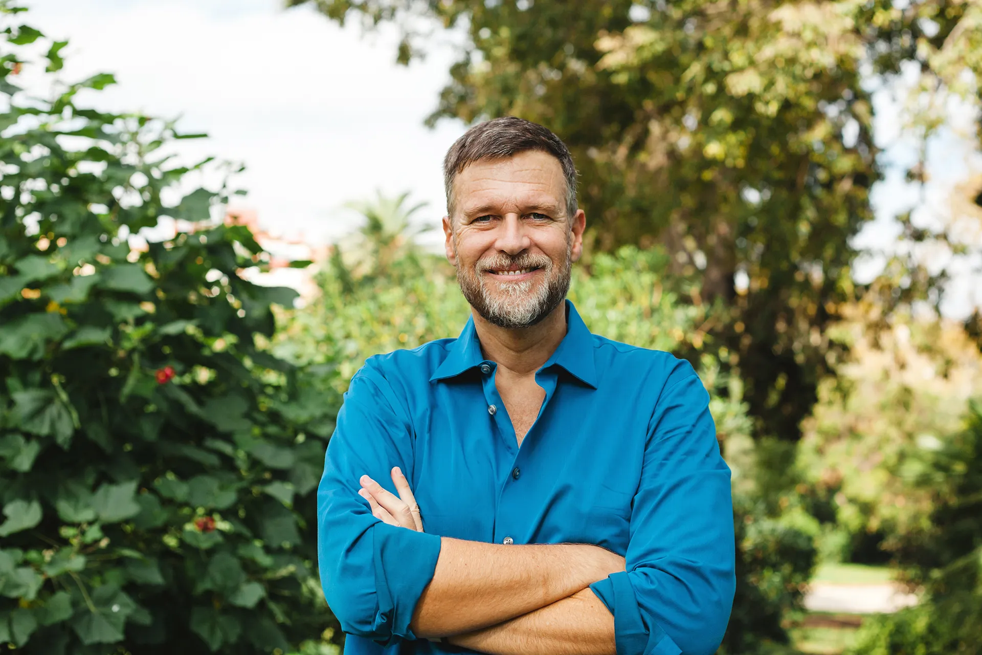 Middle-aged man with a beard smiling confidently with arms crossed, standing in a green park.
