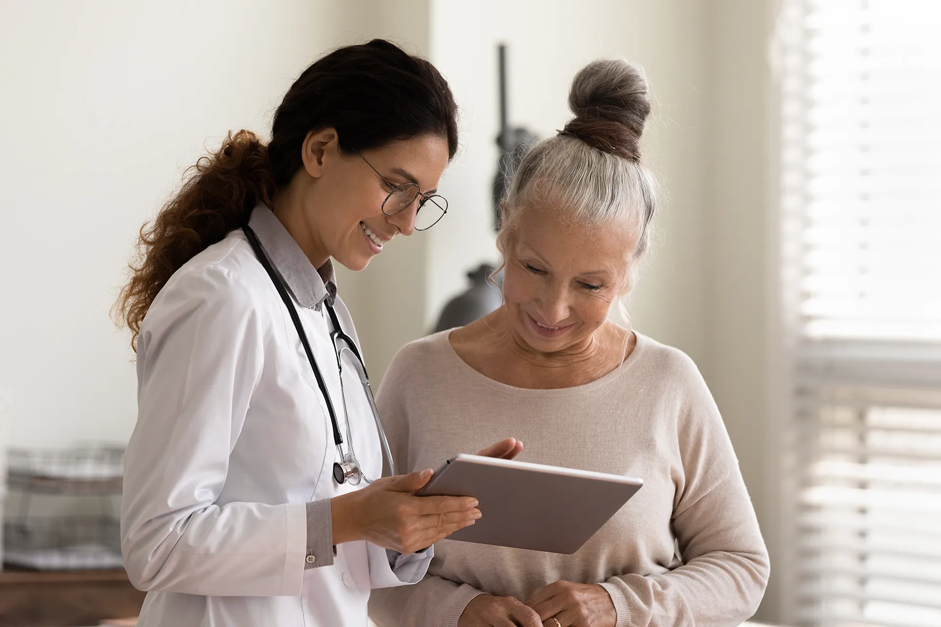 A smiling female doctor in a lab coat shows a senior woman the information displayed on a tablet, both engaged in a friendly conversation in a bright, cozy environment.
