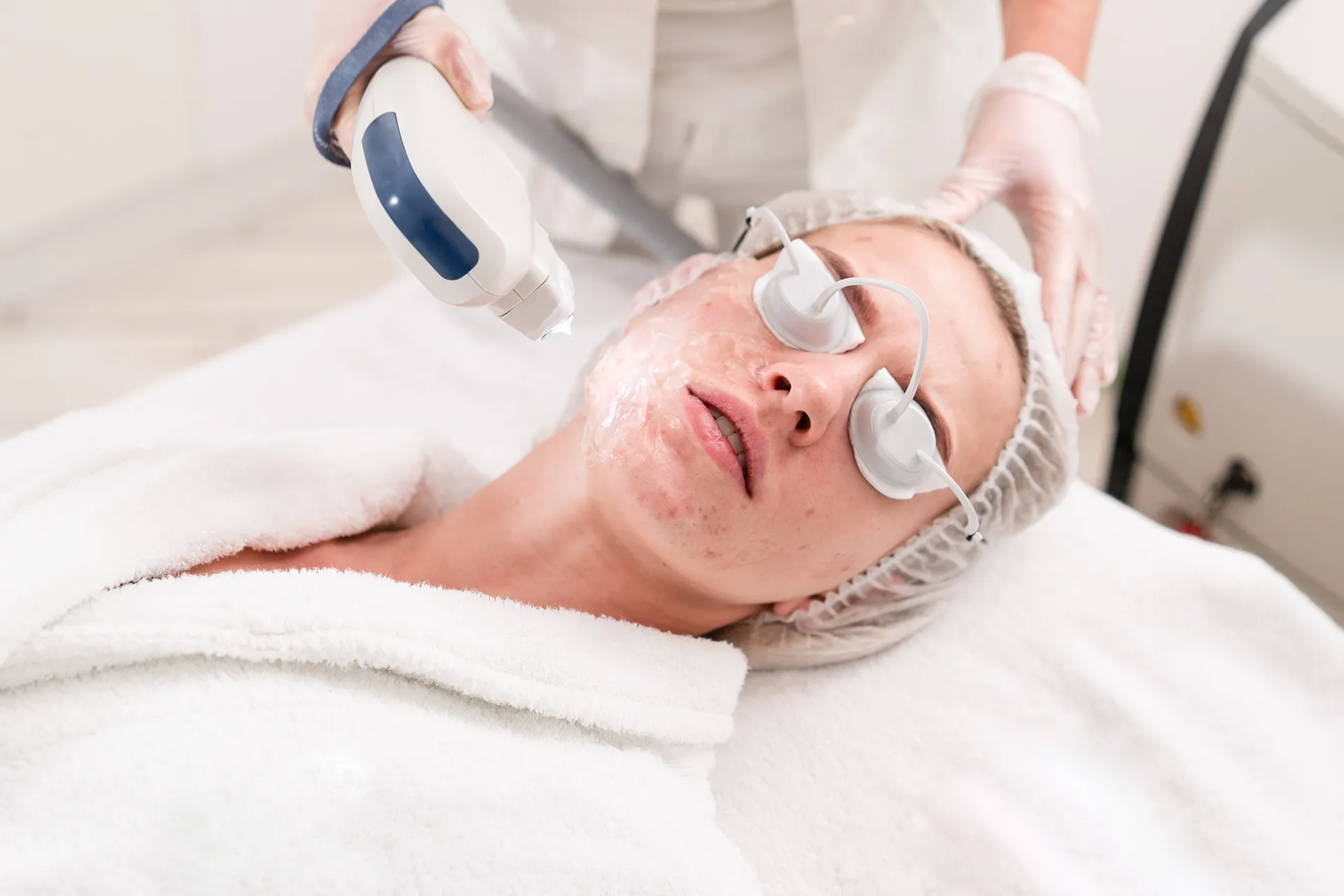 Woman receiving a facial treatment, wearing protective eyewear, with a technician using a device on her face.