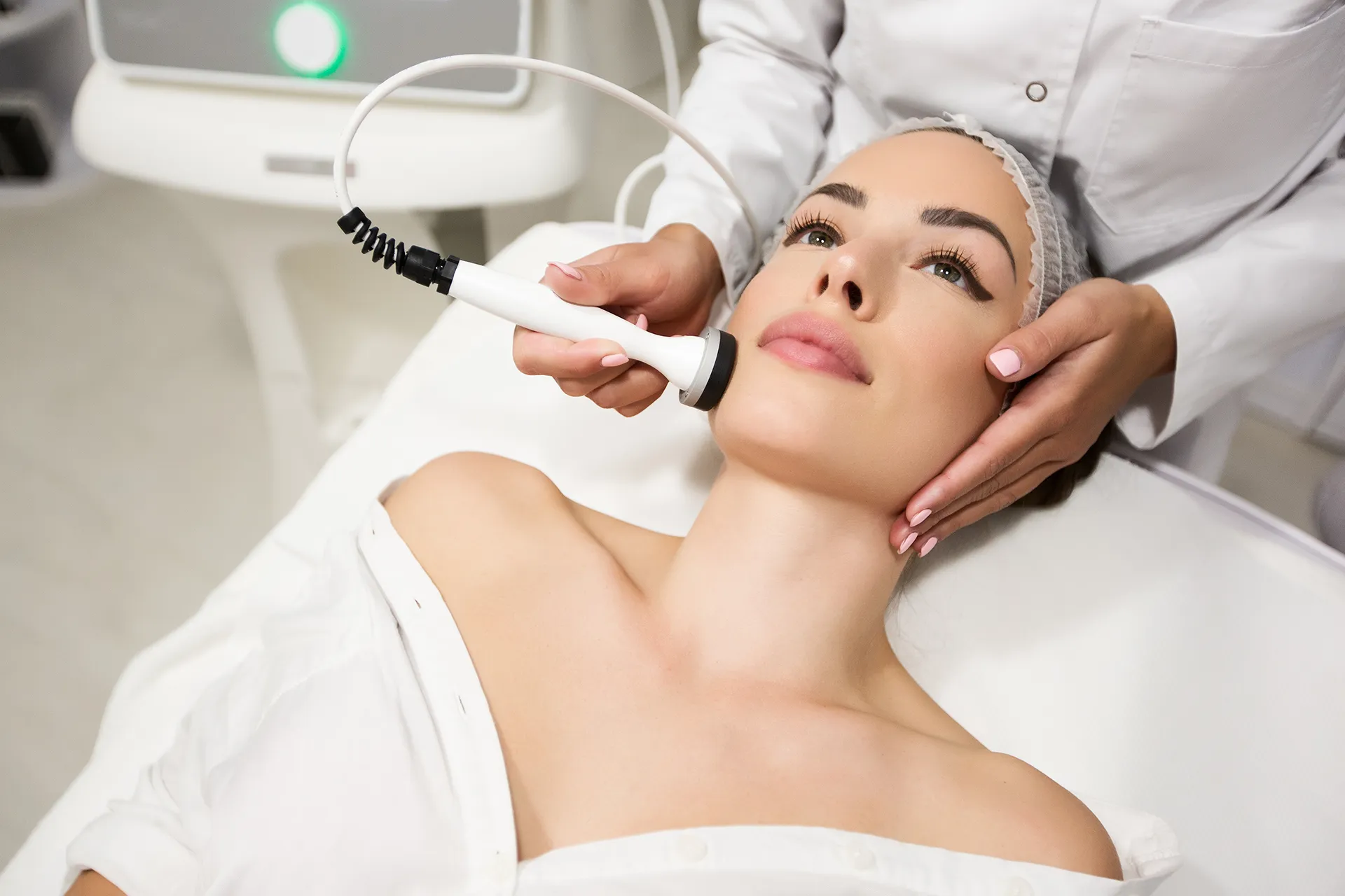 A woman lying on a treatment bed receiving a facial treatment, with a technician using a device on her face in a clinic setting.