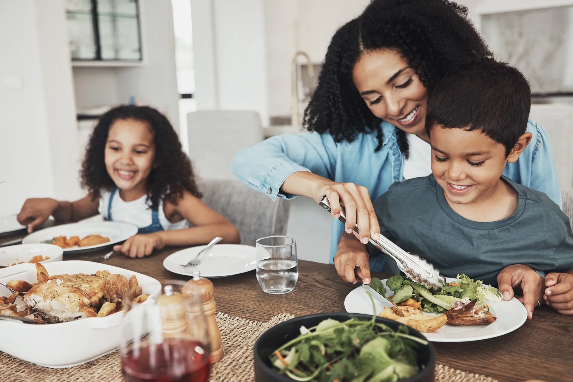 A mother serving food to her young son at a dining table, while a smiling girl sits nearby. The table is set with various dishes, including roasted chicken and salad.