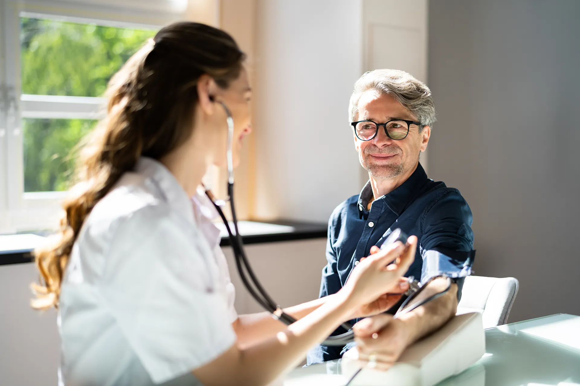 Healthcare professional taking a patient's blood pressure with a stethoscope in a bright, well-lit office.