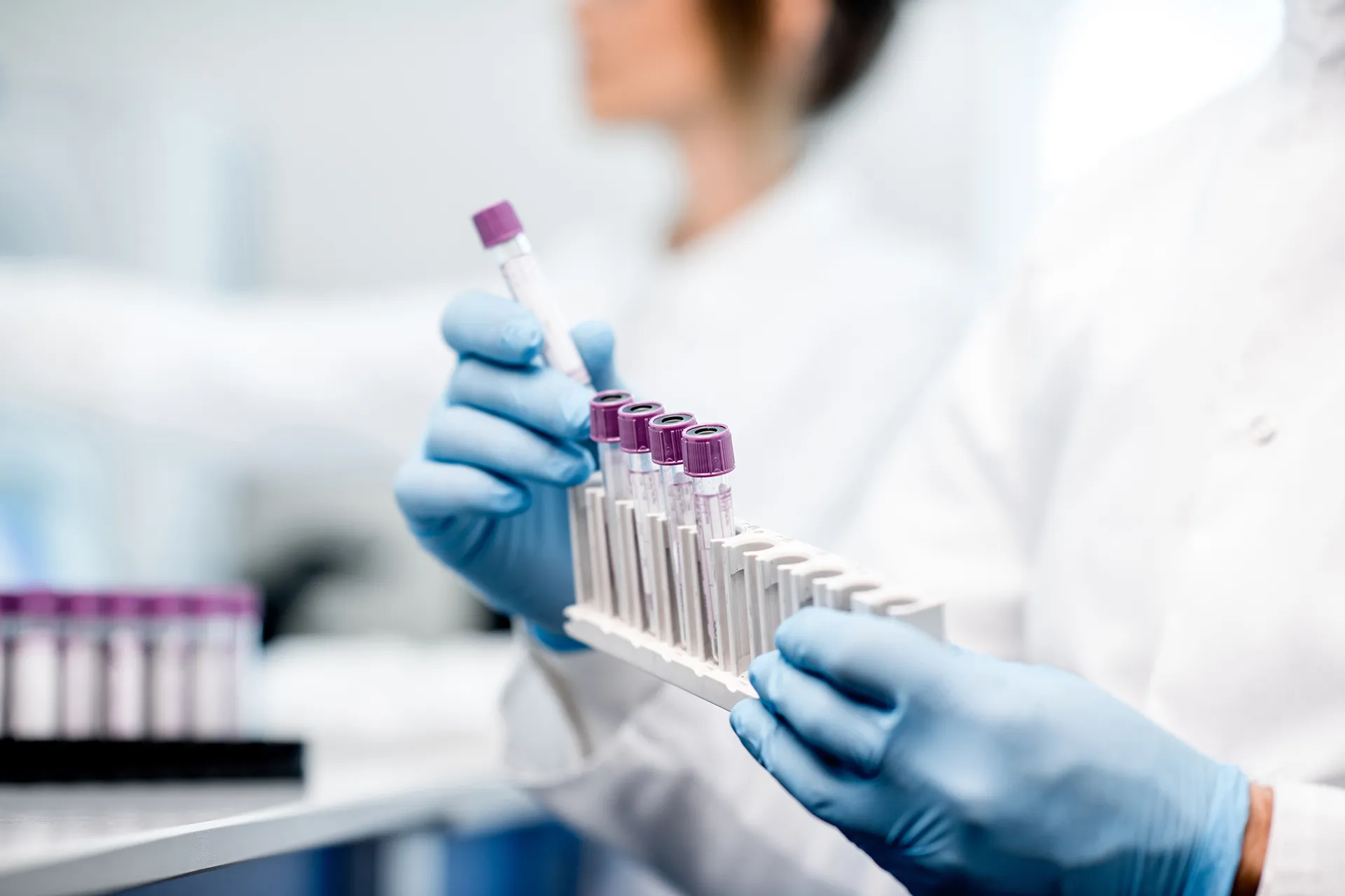 A laboratory technician holding multiple filled test tubes with purple caps in a lab setting.