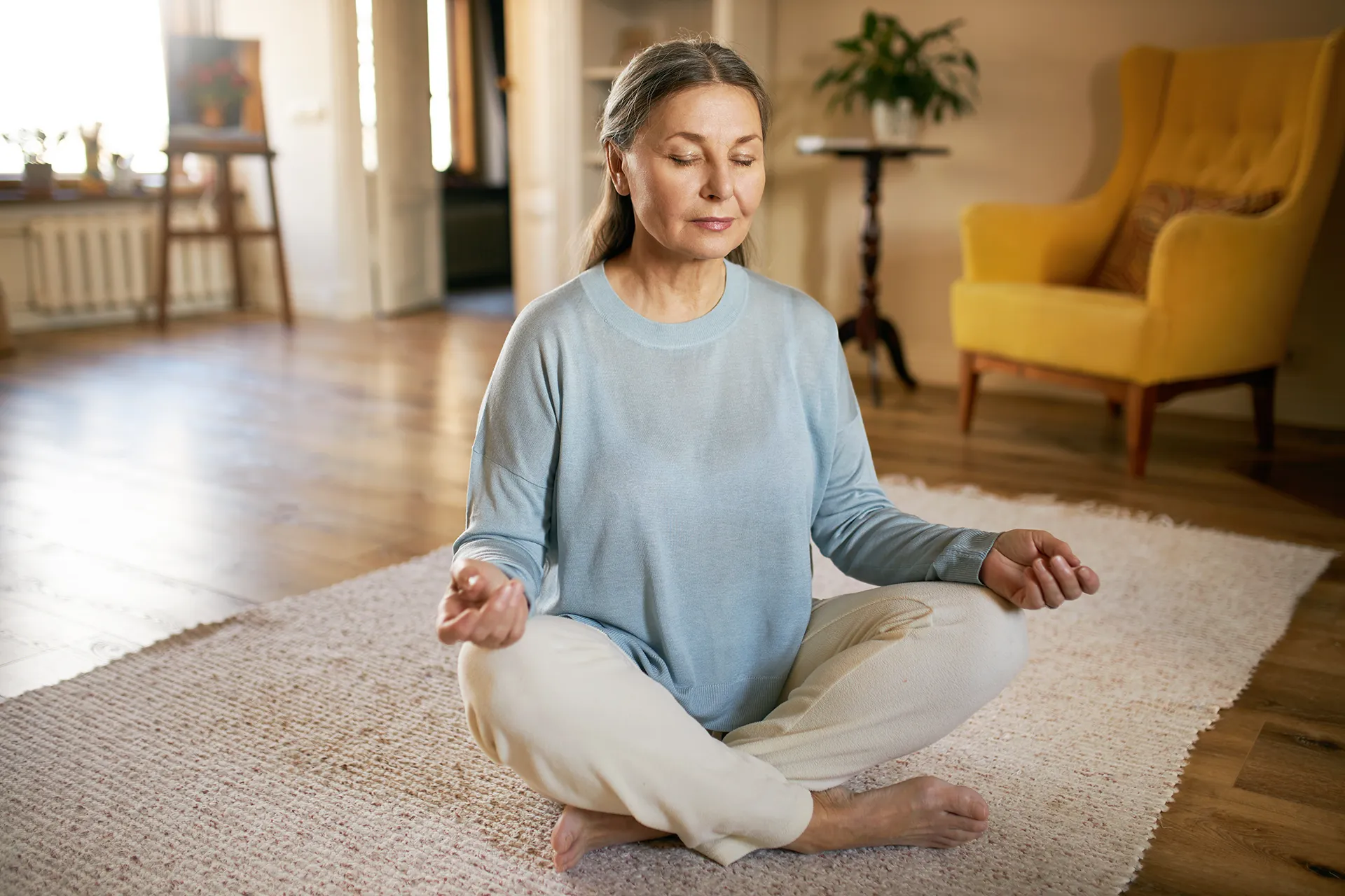 Older woman practicing meditation peacefully while sitting cross-legged on a rug indoors