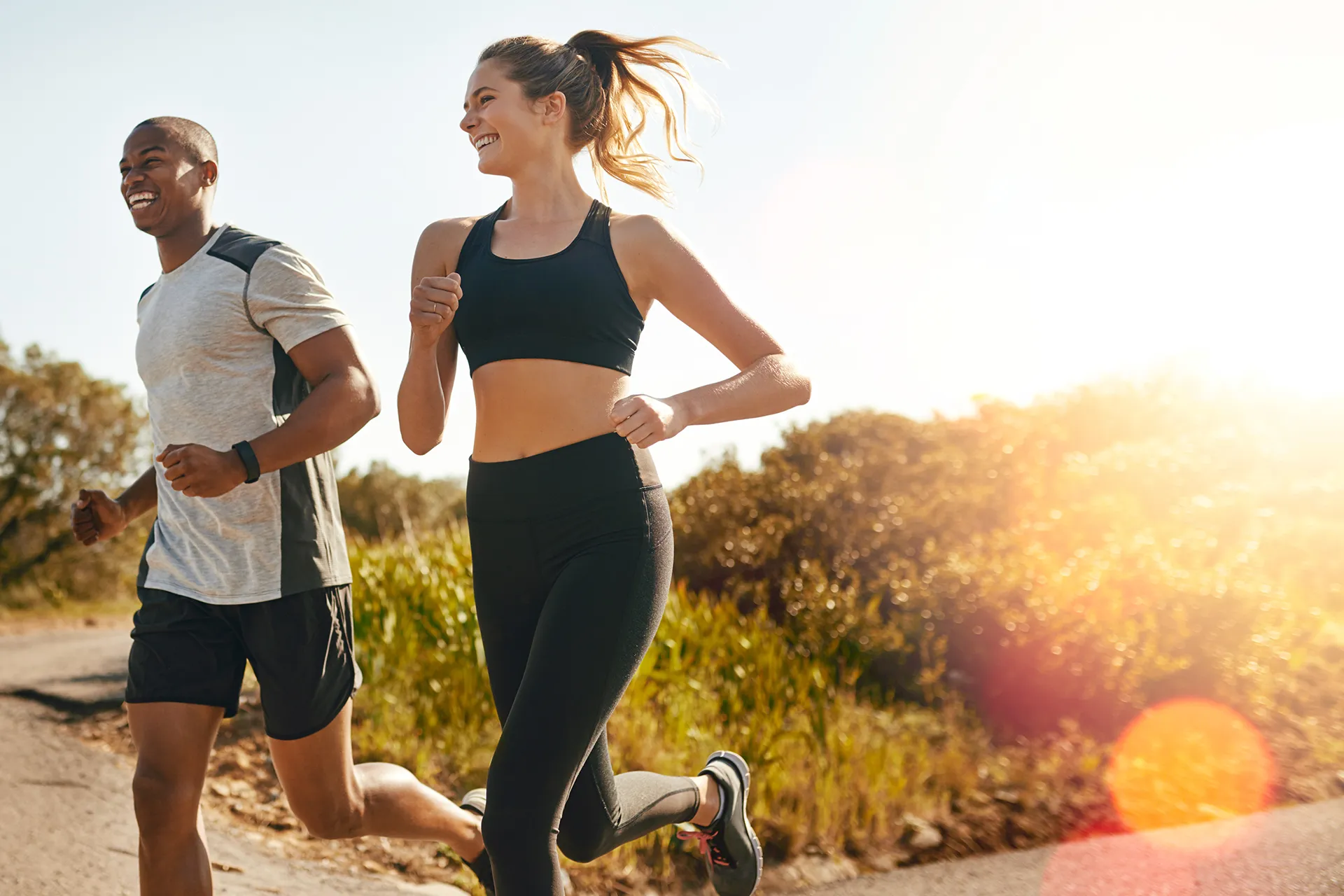 A man and woman running together outdoors along a sunlit path, both smiling and dressed in athletic wear.