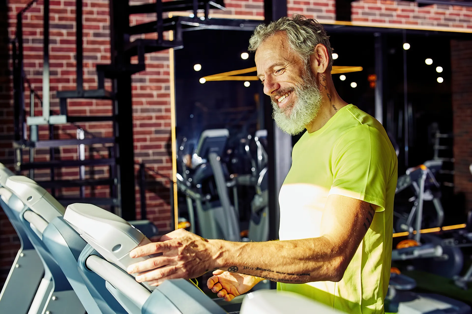 Fit older man with a beard smiling while adjusting settings on a treadmill in a modern gym.