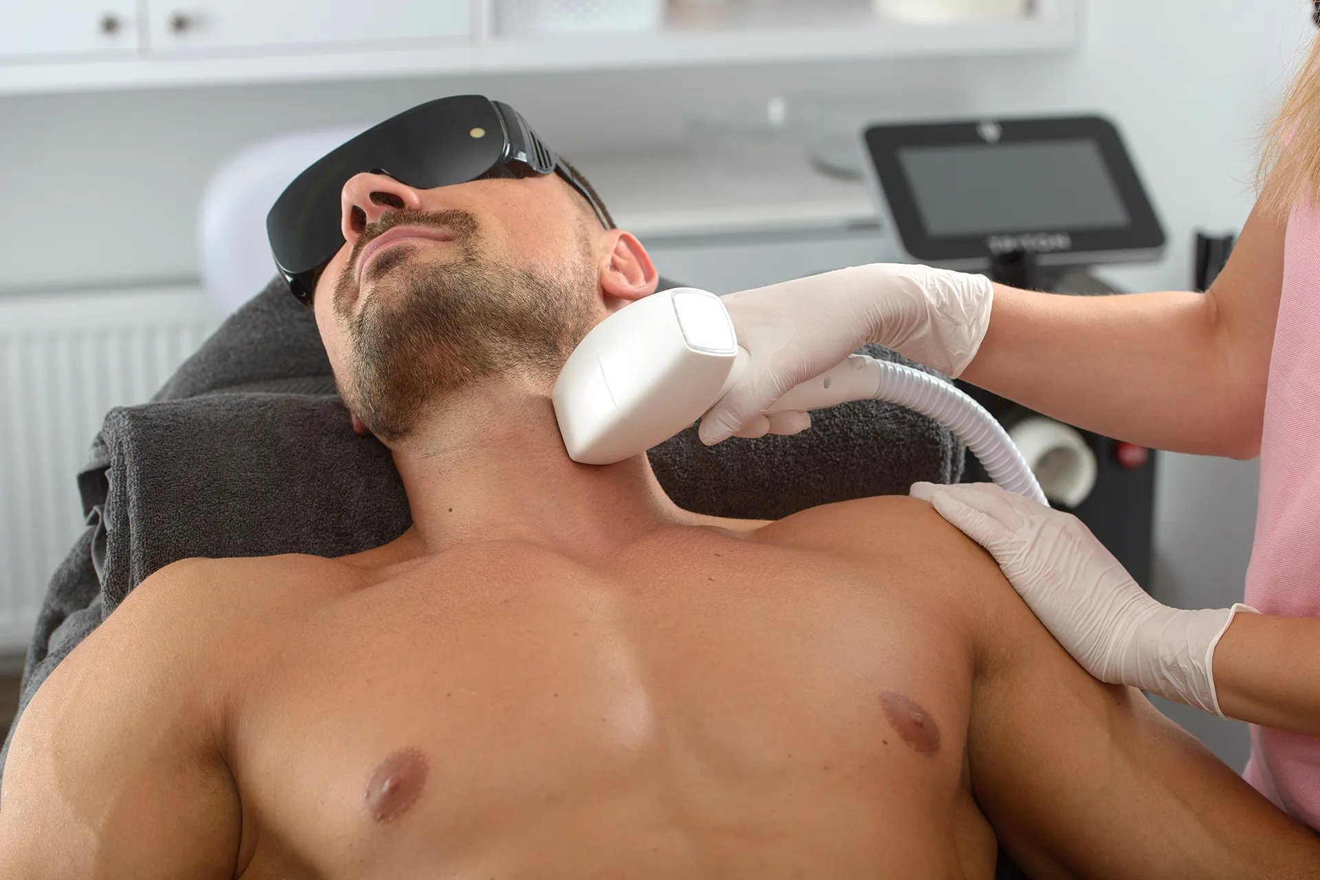 A man receiving a skin treatment in a clinic. He is lying on a treatment table, wearing protective sunglasses, while a technician operates a device on his neck.