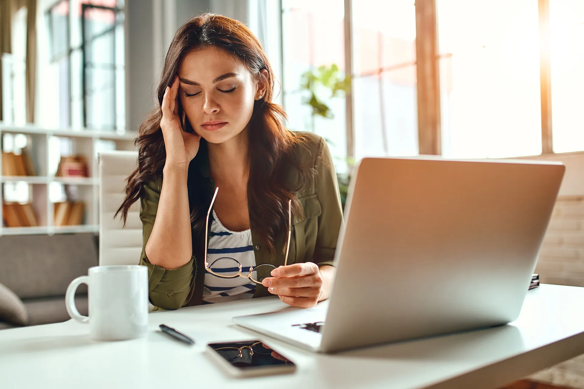 A stressed woman sitting at a desk with a laptop, holding her head in frustration, with a coffee mug and phone nearby.