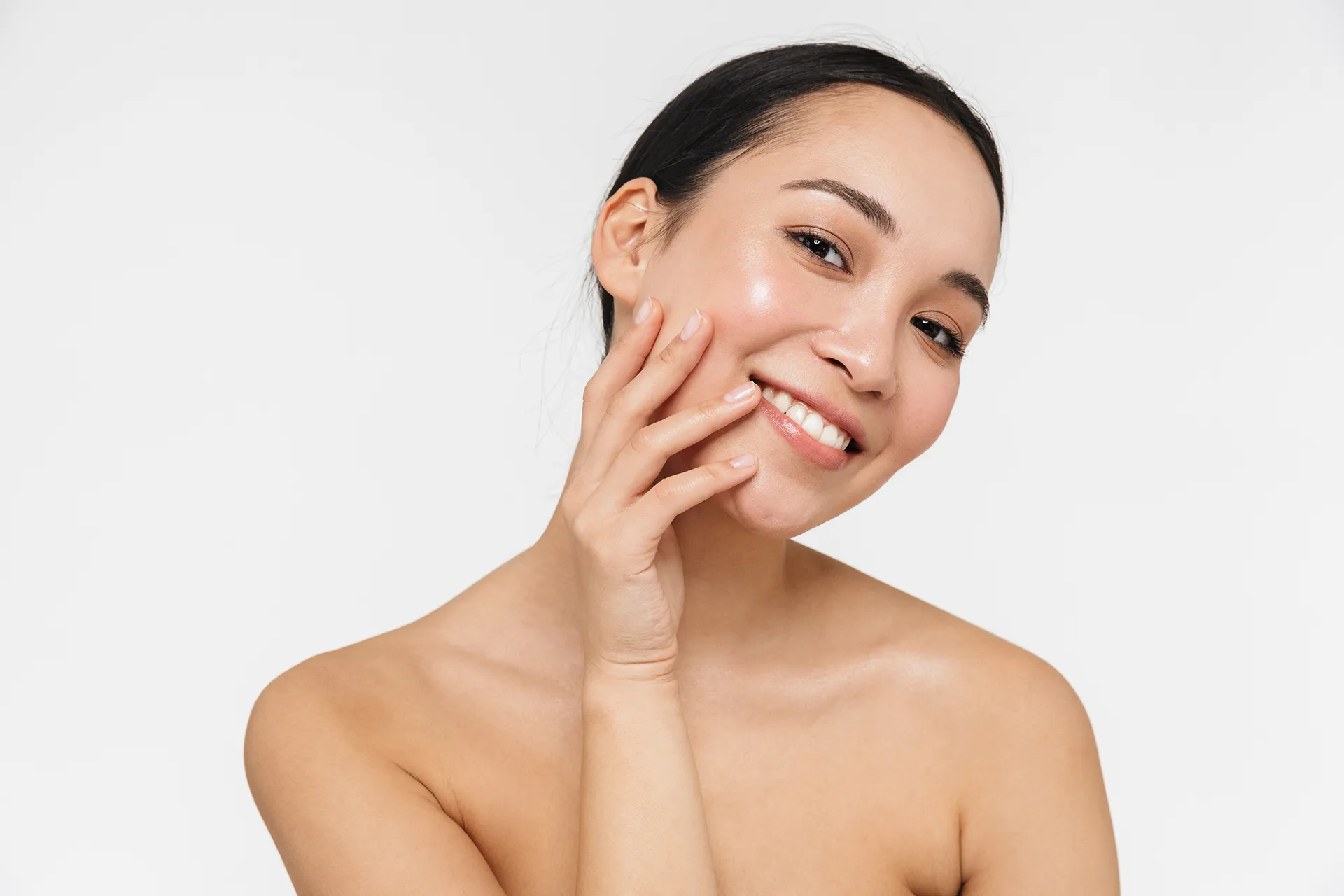 A woman with long dark hair smiles while touching her cheek, showcasing her glowing skin against a white background.